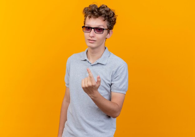 Молодой человек в черных очках, одетый в серую рубашку поло, показывает указательный палец, улыбаясь счастливым и позитивным жестом, стоящим над оранжевой стеной