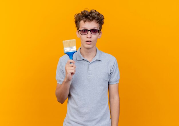 Young man in black glasses wearing grey polo shirt showing paint brush surprised over orange