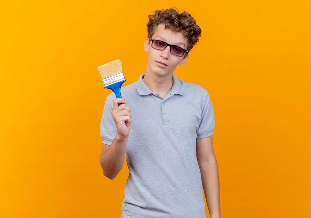 Young man in black glasses wearing grey polo shirt showing paint brush with serious face over orange