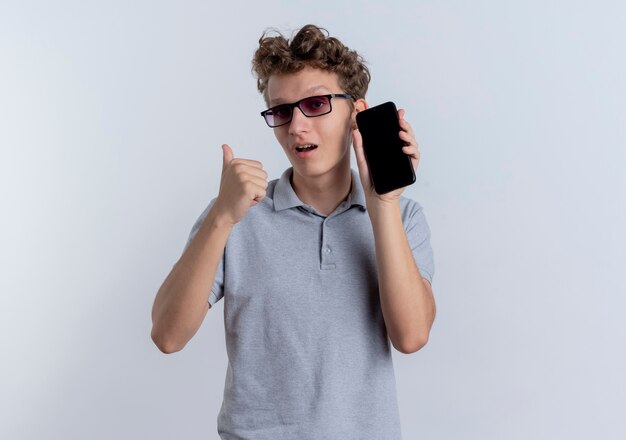 Young man in black glasses wearing grey polo shirt showing smartphone pointing back looking confused standing over white wall