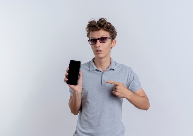 Young man in black glasses wearing grey polo shirt showing smartphone pointing with finger at it being confused standing over white wall