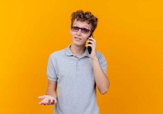 オレンジ色の手でジェスチャーを笑顔携帯電話で話している灰色のポロシャツを着た黒い眼鏡の若い男