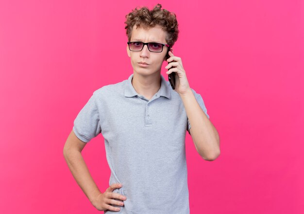 Young man in black glasses wearing grey polo shirt talking on mobile phone  with serious face standing over pink wall