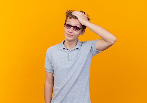 Young man in black glasses wearing grey polo shirt touching his head for mistake being confused standing over orange wall