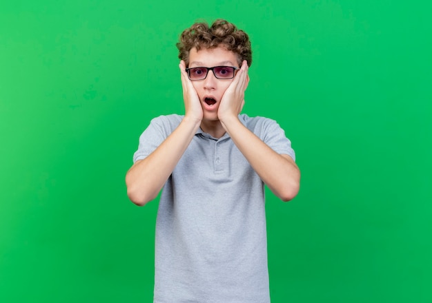 Young man in black glasses wearing grey polo shirt with hands on face being shocked over green