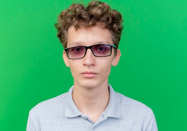 Young man in black glasses wearing grey polo shirt with serious face over green