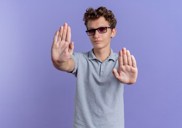 Young man in black glasses wearing grey polo shirt  with serious face making stop gesture with hands standing over blue wall