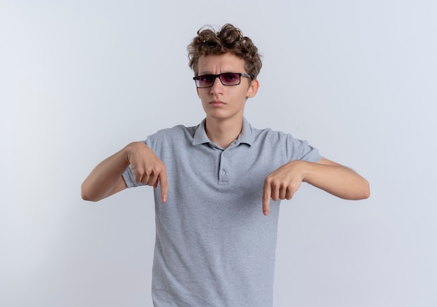 Young man in black glasses wearing grey polo shirt  with serious face pointing down with index fingers standing over white wall