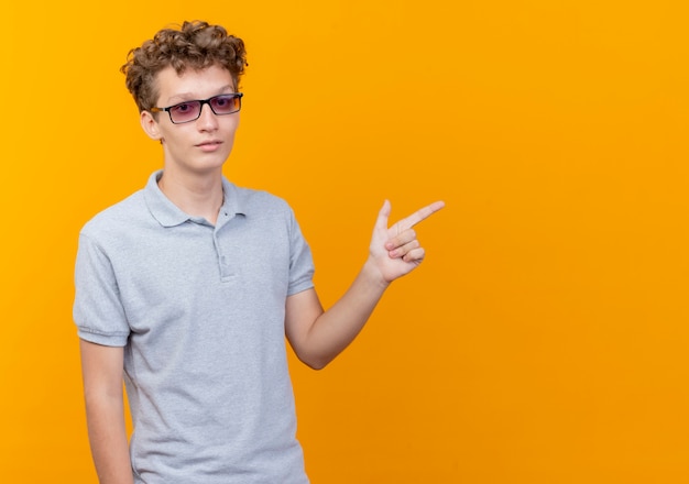Foto gratuita giovane uomo con gli occhiali neri che indossa una polo grigia con la faccia seria che punta con il dito di lato sopra l'arancio