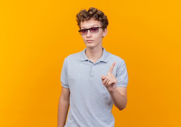 Young man in black glasses wearing grey polo shirt with serious face showing index finger warning gesture standing over orange wall