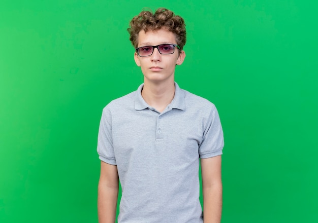 Young man in black glasses wearing grey polo shirt  with serious face standing over green wall