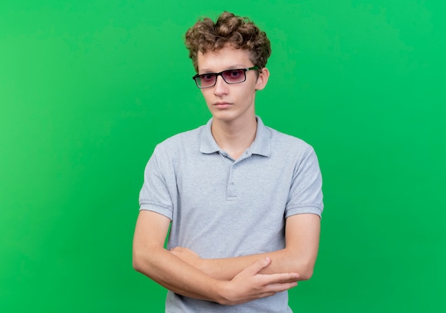 Молодой человек в черных очках в серой рубашке поло с серьезным лицом со скрещенными руками стоит над зеленой стеной