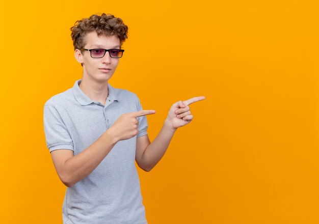 Young man in black glasses wearing grey polo shirt  with smile on face pointing with fingers to the side standing over orange wall