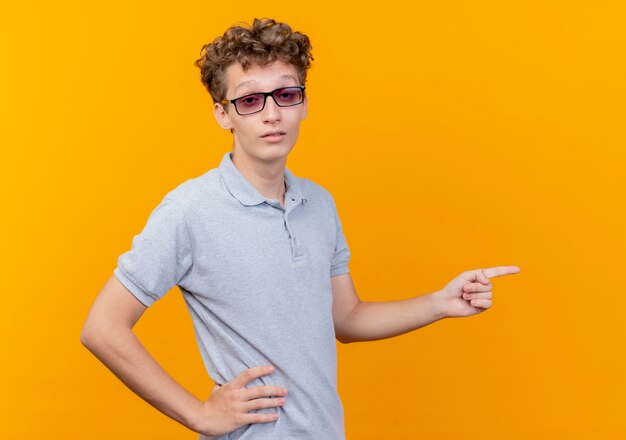 Young man in black glasses wearing grey polo shirt with smile pointing with finger to the side over orange