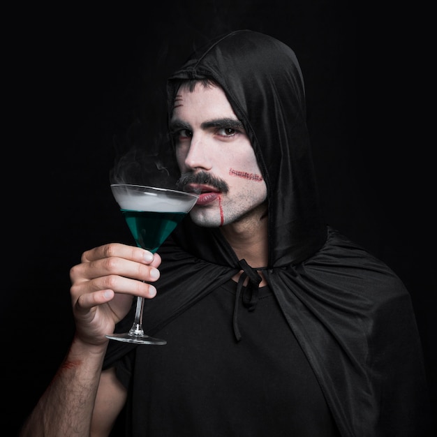 緑色の液体のガラスとスタジオでポーズを取る黒のハロウィンの外套の若い男