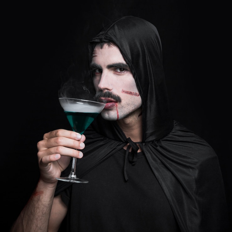 Free Photo young man in black halloween cloak posing in studio with glass of green liquid