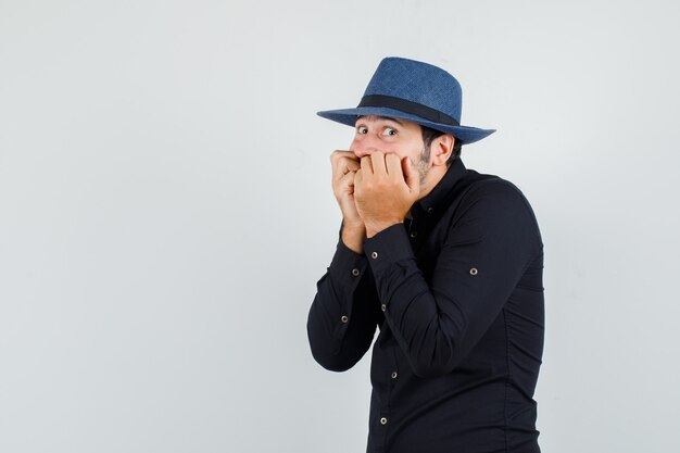 Young man in black shirt, hat covering mouth behind knuckle and looking scared.