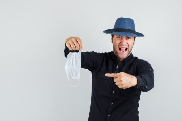 Young man in black shirt, hat pointing at his medical mask and looking excited