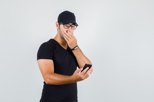 Young man in black t-shirt, cap, glasses looking at smartphone with hand on mouth and looking surprised