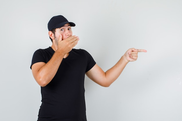 Young man in black t-shirt, cap pointing to side with hand on mouth and looking surprised