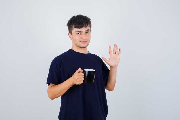 Young man in black t-shirt holding cup of tea, showing palm and looking confident , front view.