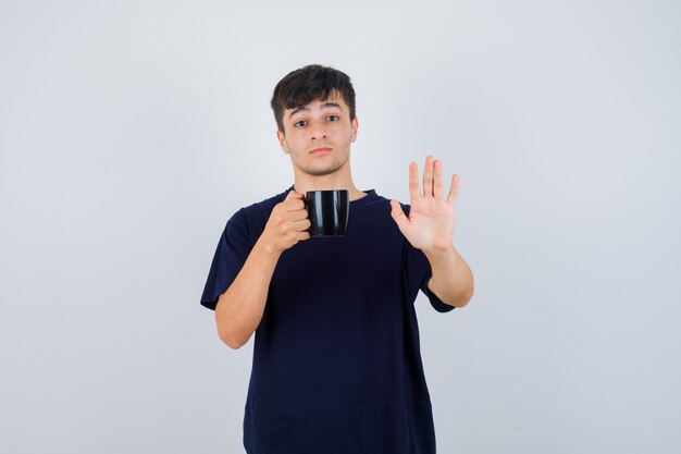 Young man in black t-shirt holding cup of tea, showing stop gesture and looking scared , front view.