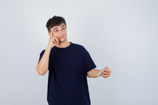 Young man in black t-shirt holding mobile phone, keeping hand on head and looking thoughtful , front view.