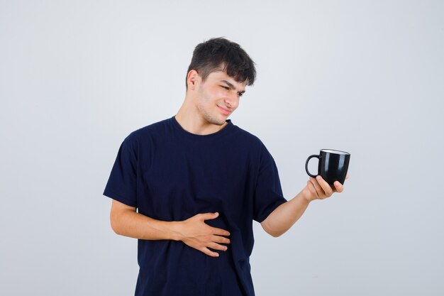 Young man in black t-shirt looking at cup and looking pensive , front view.