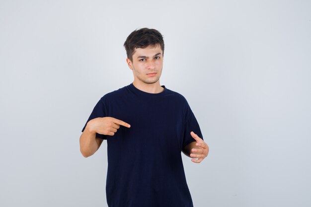 Young man in black t-shirt pointing at his palm spread out and looking serious , front view.