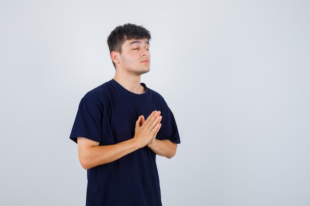 Young man in black t-shirt showing namaste gesture and looking hopeful , front view.