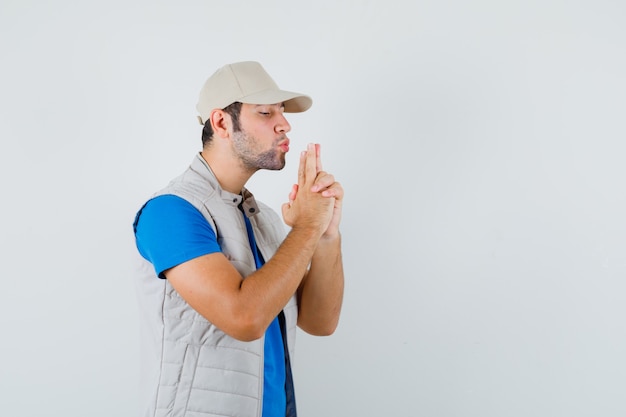 Young man blowing on finger pistol in t-shirt, jacket, cap and looking confident. front view.