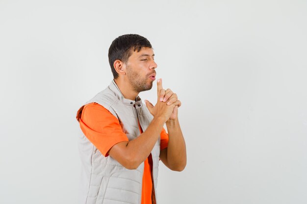 Young man blowing on gun made by fingers in t-shirt, jacket and looking confident , front view.