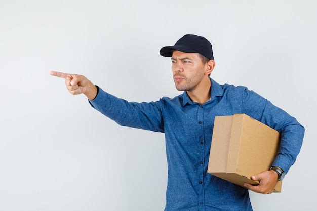 Free photo young man in blue shirt, cap holding cardboard box while pointing away , front view.