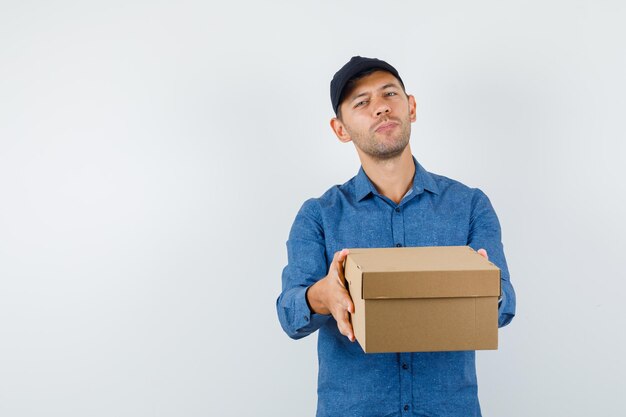 Young man in blue shirt, cap presenting cardboard box , front view.