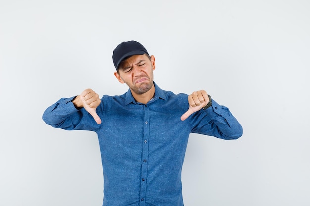 Free photo young man in blue shirt, cap showing thumbs down and looking disappointed , front view.