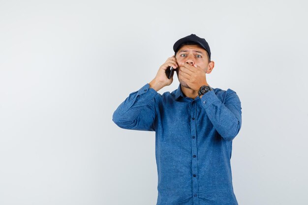 Молодой человек в голубой рубашке, кепке разговаривает по мобильному телефону и выглядит удивленным, вид спереди.