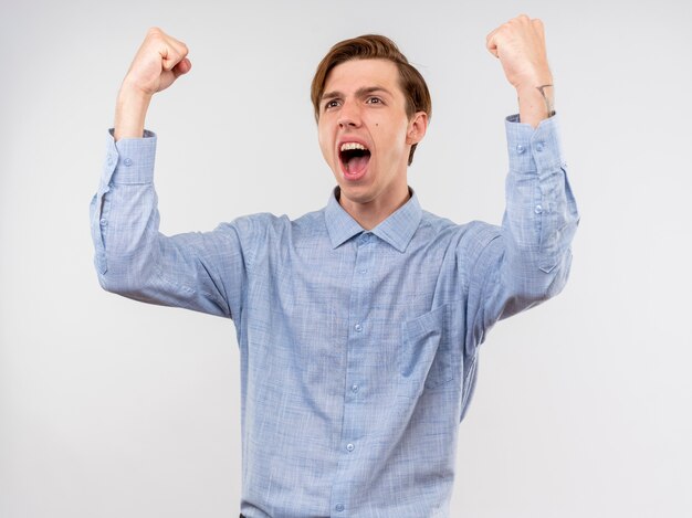 Young man in blue shirt clenching fists happy and excited rejoicing his success standing over white wall