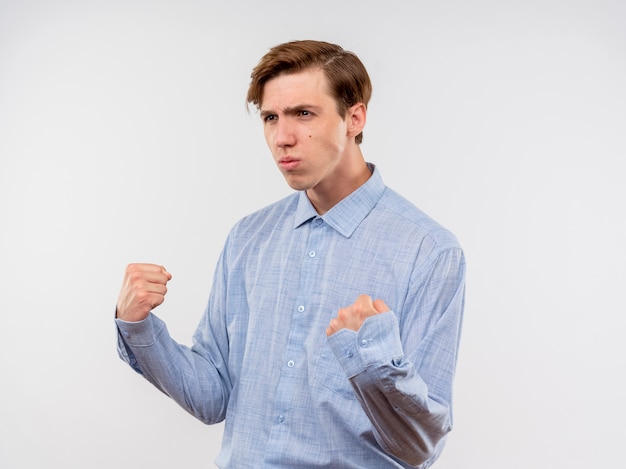 Young man in blue shirt clenching fists looking confident standing over white wall