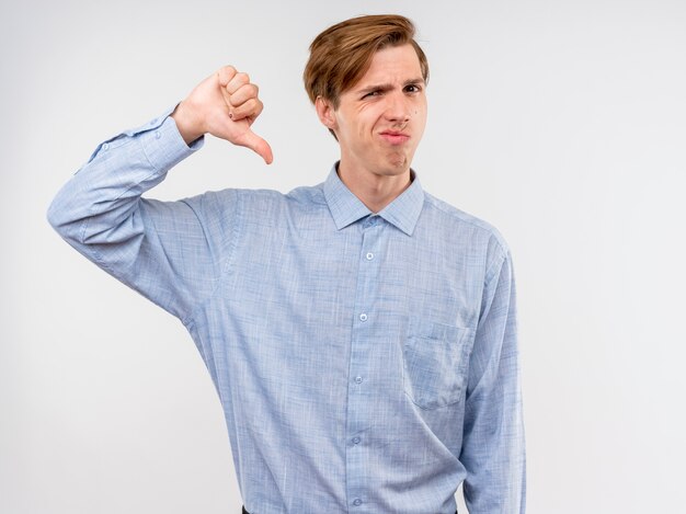 Young man in blue shirt  displeased showing thumbs down standing over white wall