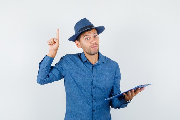 Young man in blue shirt, hat holding clipboard with finger up and looking curious , front view.