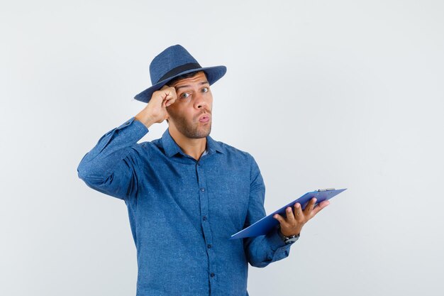 Young man in blue shirt, hat holding clipboard with hand on temples and looking thoughtful , front view.