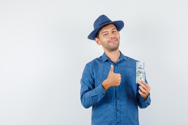 Young man in blue shirt, hat holding dollar banknote with thumb up and looking merry , front view.