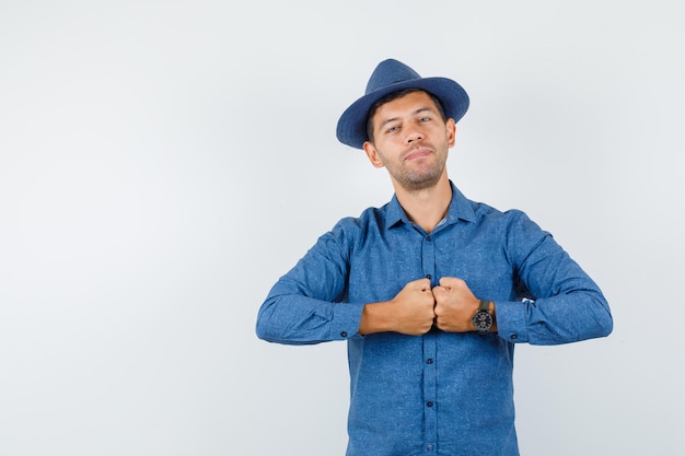 Giovane in camicia blu, cappello che tiene i pugni e sembra fiducioso, vista frontale.