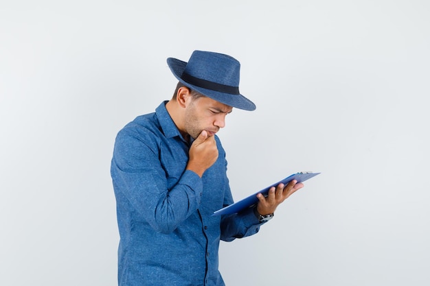 Young man in blue shirt, hat looking over notes on clipboard and looking busy , front view.