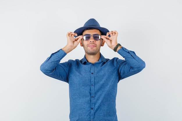 Young man in blue shirt, hat touching his glasses and looking cheerful , front view.