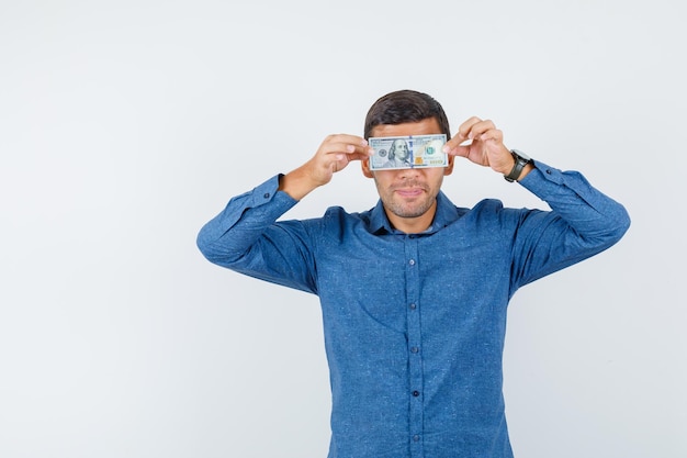 Young man in blue shirt holding dollar bill on eyes and looking funny , front view.