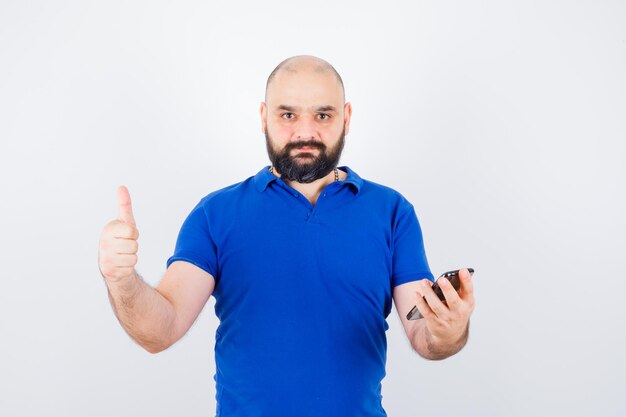 Young man in blue shirt holding phone while showing thumb up , front view.