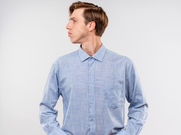 Young man in blue shirt lookign aside with serious confident expression standing over white wall