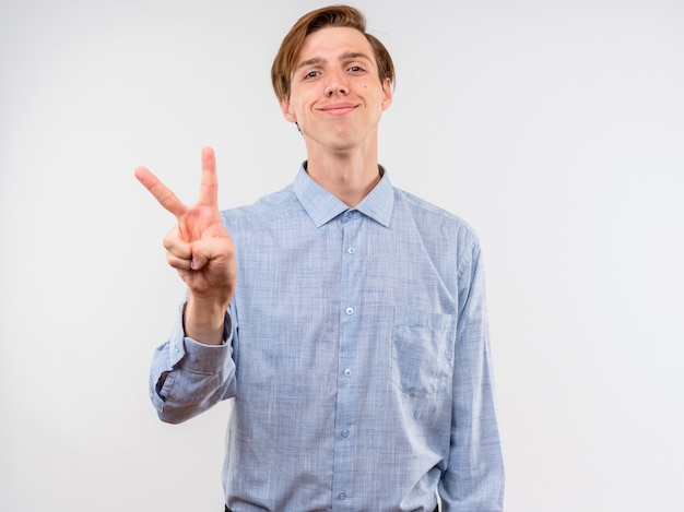 Young man in blue shirt lookign at camera smiling with happy face showing v-sign standing over white background