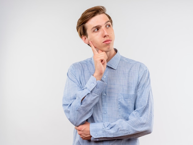 Young man in blue shirt looking aside with pensive expression thinking standing over white background